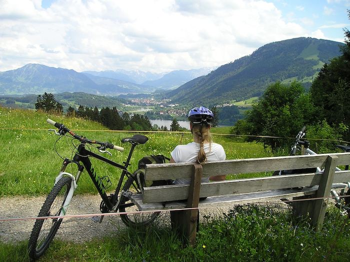 Mädchen sitzt auf einer Bank mit angelehntem Fahrrad und betrachtet eine Berglandschaft