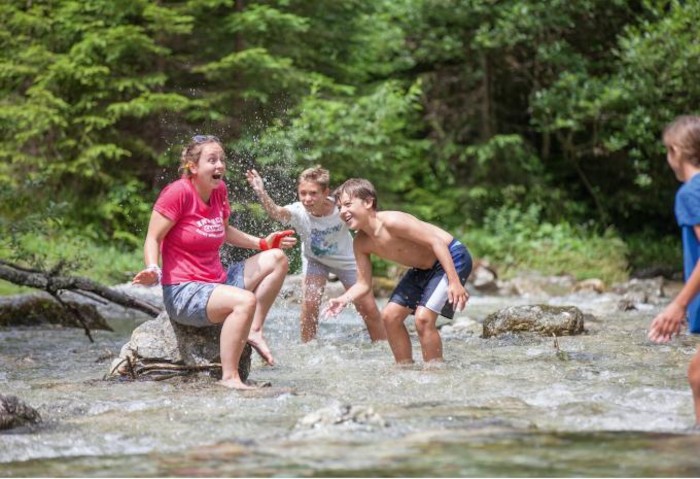 Zwei Kinder spritzen eine Betreuerin im Fluss nass im Ferienlager Waadt.