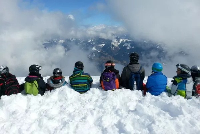 Kinder und Jugendliche sitzen auf einem Berg in einer Reihe im Schnee und betrachten die Gebirgskulisse des Salzburger Alpenlands