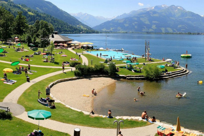 Feriengäste entspannen sich bei strahlendem Sonnenschein in einem dicht begrünten Freibad am Ufer des Zeller Sees mit dem Salzburger Alpenland im Hintergrund