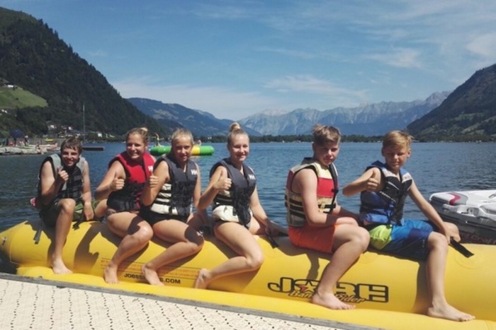 Junge Feriengäste posieren auf einer Wassersport-Banane am Ufer des sommerlichen Zeller Sees mit dem Salzburger Alpenland im Hintergrund