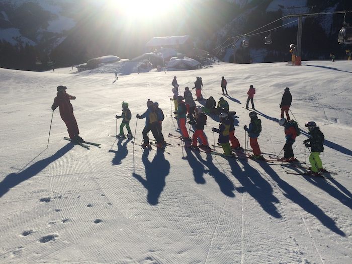 Viele Kinder stehen im Winterferienlager vor einem Skilehrer und hören Ihm zu