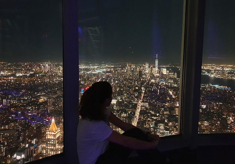 Ein Mädchen sitzt am Fenster und blickt über die hell erleuchtete Stadt New York bei Nacht bei unseren Sprachreisen USA für Schüler in Gastfamilie