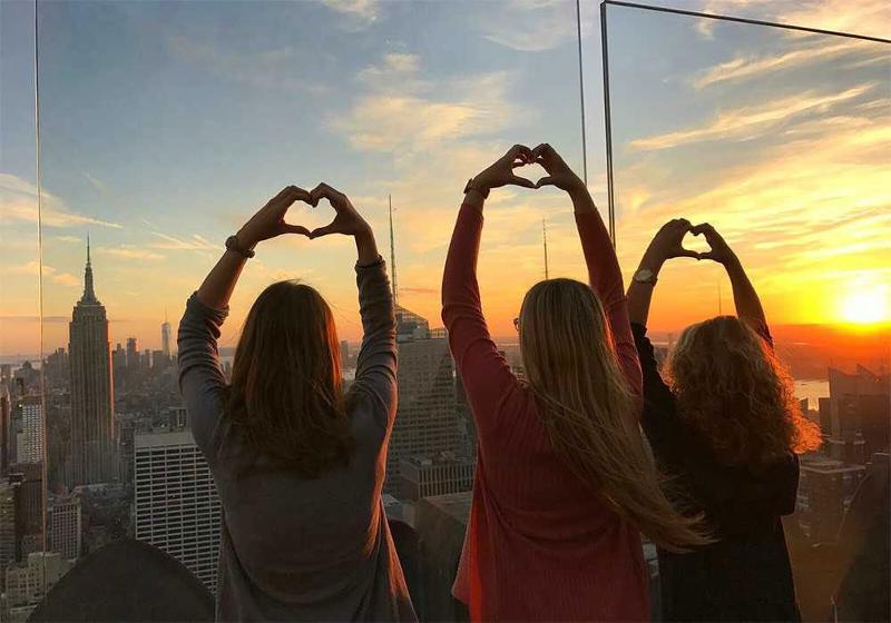  Drei Mädchen posieren gemeinsam auf einem Dachgeschoss beim Sonnenuntergang und halten die Hände zu Herzen geformt in die Luft während unserer Sprachreisen New York für Schüler