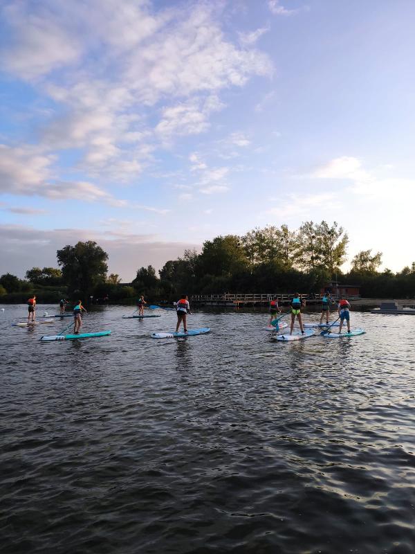 Man sieht die Natur des Ferienlager Vorpommern und Kinder die alle zusammen auf ihren Stand up Paddles stehen und Spaß zusammen auf dem See haben und das wunderschöne Wetter genießen 