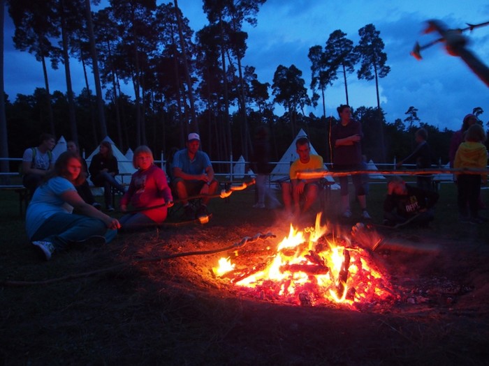 Einige Kinder sitzen bei Sonnendämmerung draußen vor ihren Zelten vor einem großen Lagerfeuer und halten ihr Stockbrot über die großen Flammen des Lagerfeuers bei dem Zeltlager Pfingsten