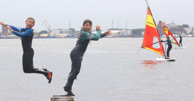 Im Hintergrund stehen Kids auf dem Surfbrett mit dem Segel in der Hand während vom Steg zwei Kinder im Windsurfcamp ins Wasser springen.