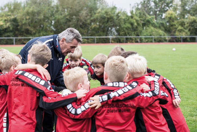 Dieter Müller steht in einem Kreis umringt von Kindern und erklärt Fußball-Theorie!