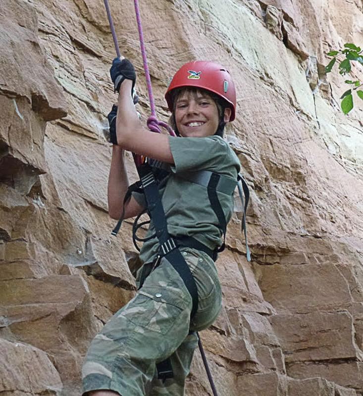 Ein Junge hängt im Abenteuercamp mit Seilen gesichert an einer Felswand.
