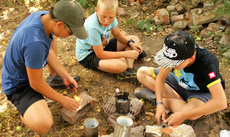 Drei Jungs probieren im Abenteuercamp über ihrem selbst entfachtem Feuer zu kochen.