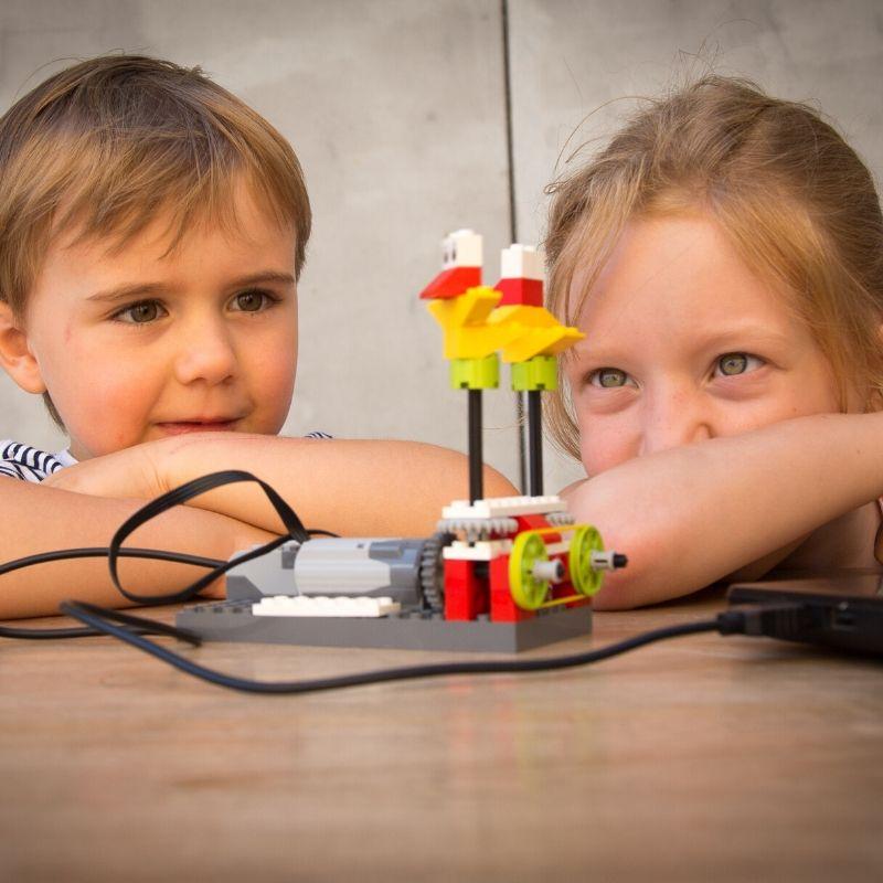 Dois jovens estão fascinados a observar uma construção de LEGO que se mexe, interagindo com um motor ligado a um portátil, simbolizando a diversão e a criatividade que podem surgir nos cursos de programação para jovens.