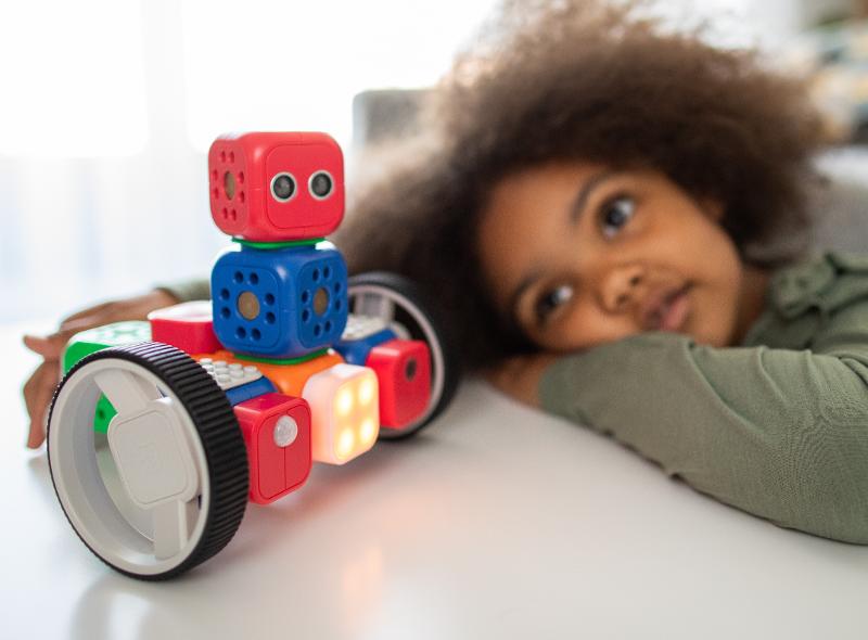 Uma criança observa atentamente um robô colorido e divertido, feito de blocos, que se destaca numa mesa, simbolizando a criatividade e a descoberta que os cursos de programação para jovens podem oferecer.