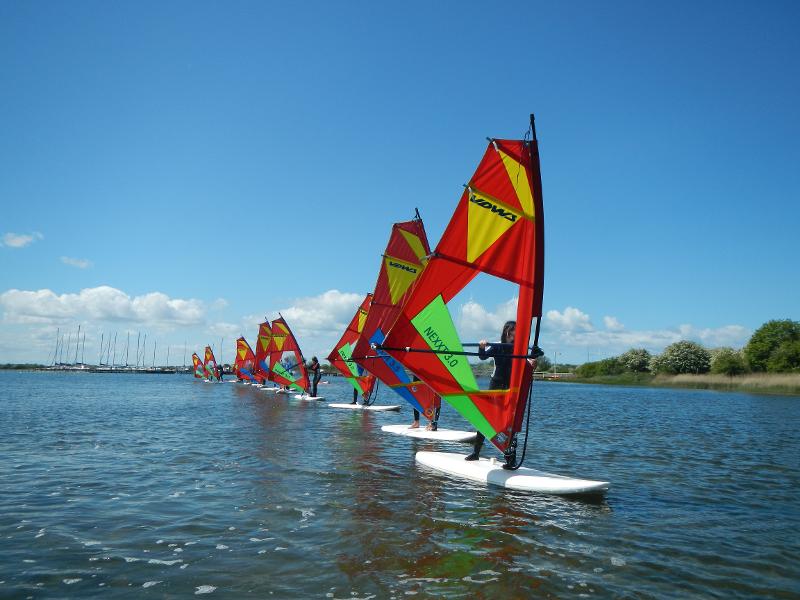 Die Kids fahren im Windsurfcamp in einer geraden Linie hintereinander auf ihren Boards über das Wasser.