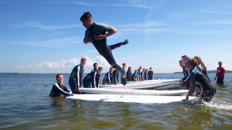 Bei einer lustigen Übung im Windsurfcamp springt ein Junge von den Boards ins Wasser.