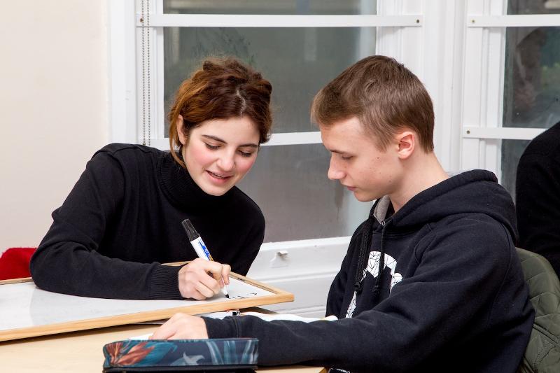 Eine Lehrerin schreibt mit einem Stift auf Englisch Sprachreise etwas auf eine Tafel während ein Schüler ihr zuschaut